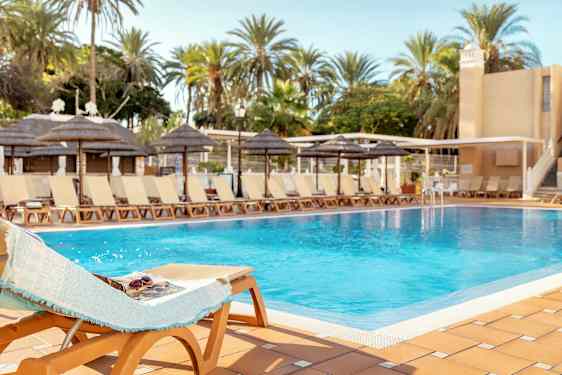 Et basseng omgitt av solsenger og palmer under en klar himmel. (AI-generert tekst)