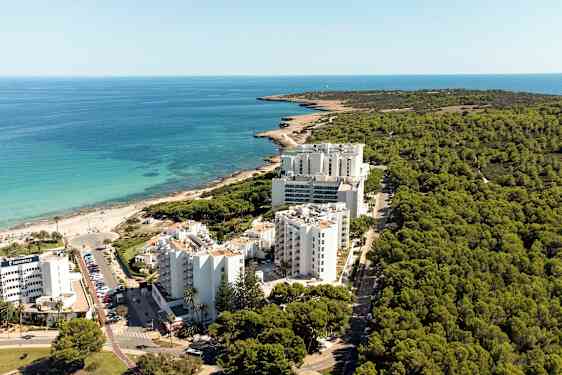 Luftfoto av en kystlinje med hotellbygninger foran en skog og nær en strand. (AI-generert tekst)