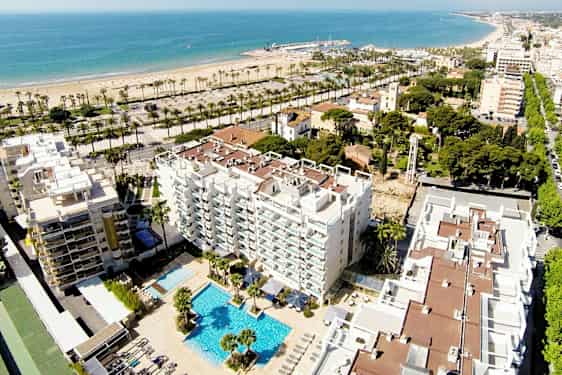 Luftfoto av et hotell med bassengområde, omgitt av bygninger og grøntarealer. I bakgrunnen ses en sandstrand og havet. (AI-generert tekst)