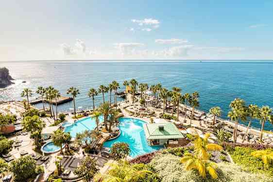 Et stort basseng med omkringliggende palmer og solsenger, plassert ved stranden med utsikt over det glitrende havet. (AI-generert tekst)