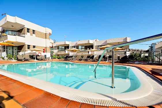 Et utendørsbasseng omgitt av solsenger og parasoller ved en bygning med flere etasjer. (AI-generert tekst)