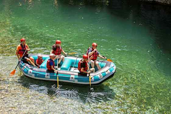 En gruppe mennesker driver med rafting i en oppblåsbar flåte på klart vann, og har på seg hjelmer og redningsvester. (AI-generert tekst)