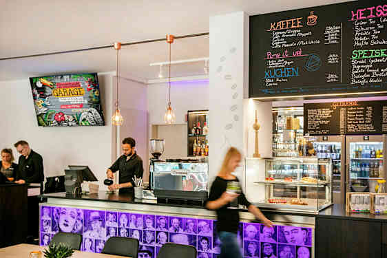 En kaféinteriør med en disk som inneholder kaffemaskin, bakverk i en glassmonter og en meny på veggen over. Ansatte står bak disken. (AI-generert tekst)