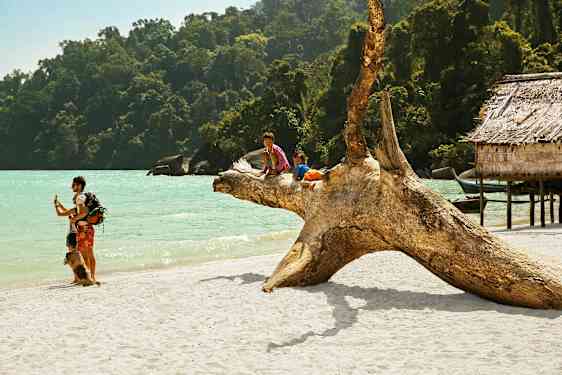 To barn sitter på en stor trestokk på en sandstrand ved havet, omgitt av frodig skog og trebygninger. (AI-generert tekst)