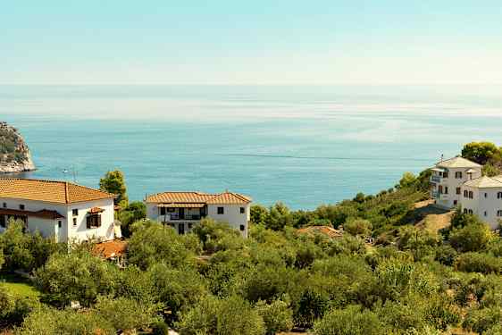 Et landskap med hvite hus og røde tak omgitt av grønn vegetasjon og utsikt over et rolig blått hav. (AI-generert tekst)