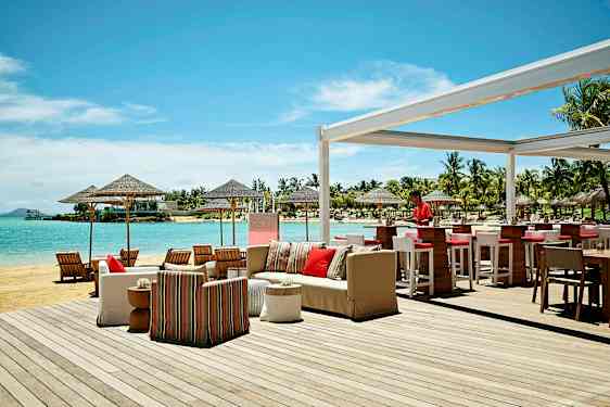 Utendørs serveringsområde ved stranden med bar og sittegrupper, samt utsikt over havet og omkringliggende palmer. (AI-generert tekst)