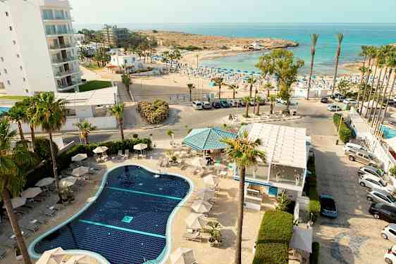 Et oversikt av et hotellbasseng med solsenger, omgitt av palmer, med havet og en solfylt strand i bakgrunnen. (AI-generert tekst)