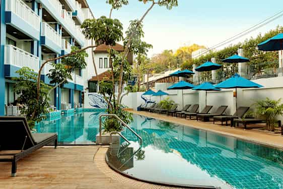 Et utendørs svømmebasseng med solsenger og parasoller, omgitt av vegetasjon og en hotellbygning med flere etasjer. (AI-generert tekst)