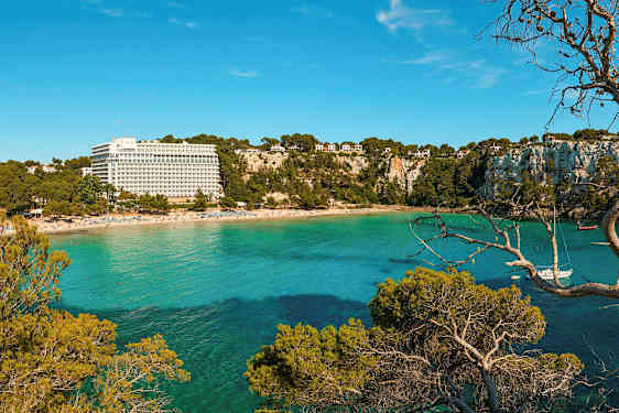 En strand med et resortbygg og frodige klipper ved et turkis hav. (AI-generert tekst)