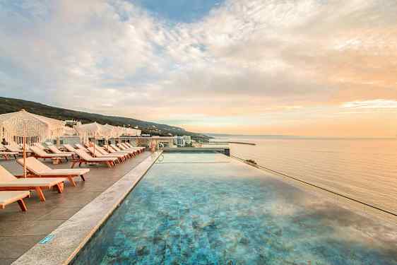 Utsikt over et infinitybasseng med solsenger og parasoller ved siden av, mot havet i solnedgang. (AI-generert tekst)