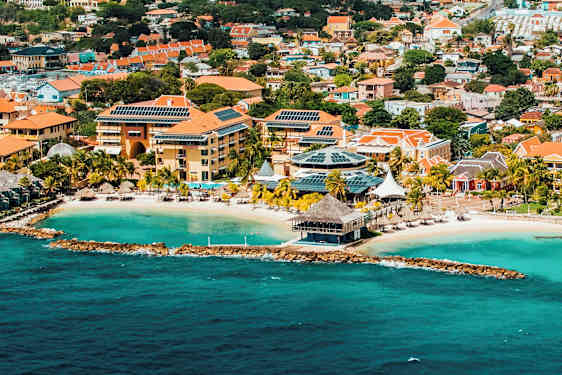 Flyfoto av en kystby med turkist vann, en sandstrand og bygninger med røde tak og solcellepaneler. (AI-generert tekst)