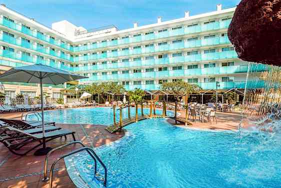 Et utendørs svømmebasseng ved en hotellbygning med solsenger, bord og parasoller. En liten bro over bassenget og en del av en fossestruktur er synlig. (AI-generert tekst)