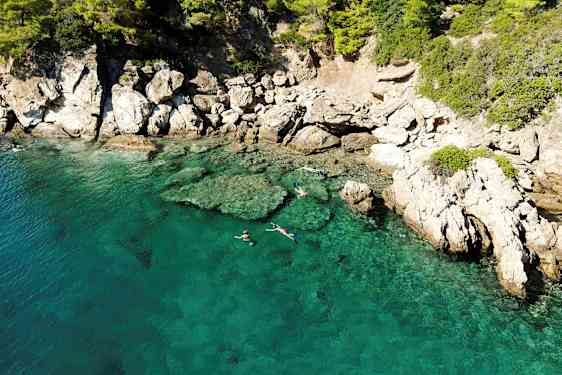Klart turkisblått vann ved en steinete kyst, hvor folk snorkler nær overflaten. (AI-generert tekst)