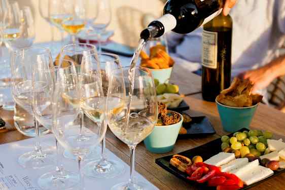 Et bord med glass fylt med hvitvin som serveres, omgitt av ulike snacks som ost, brød og frukt. (AI-generert tekst)