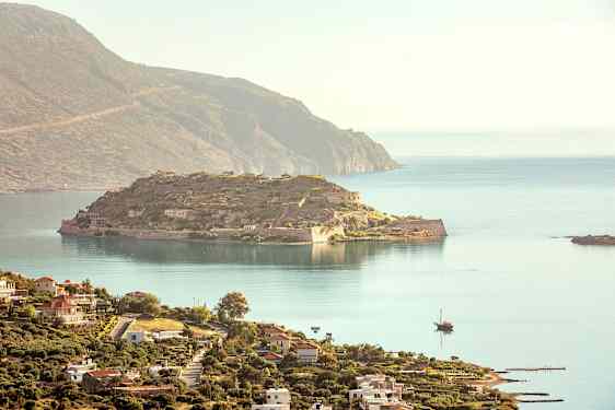 En utsikt over en historisk øy med ruiner og omkringliggende hav, og i forgrunnen sees en kystby med bygninger. (AI-generert tekst)