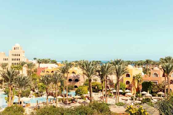 Et feriested med gule og oransje bygninger, omgitt av palmer og grønt, med bassenger og parasoller. (AI-generert tekst)