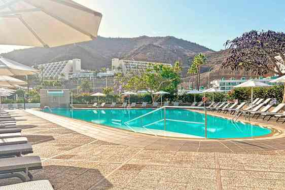 Et utendørs basseng med solsenger og parasoller, omgitt av grønne områder og fjell. (AI-generert tekst)