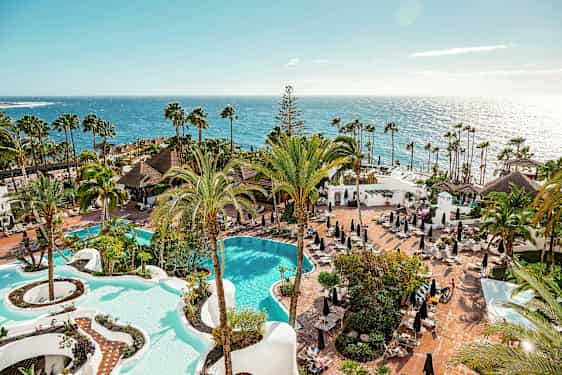 Utsikt over et hotellområde med to bassenger, palmer og solsenger ved havet. (AI-generert tekst)