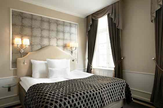Et elegant hotellrom med en dobbeltseng, dekorative puter og smakfull innredning. Gardiner rammer inn et lyst vindu. (AI-generert tekst)