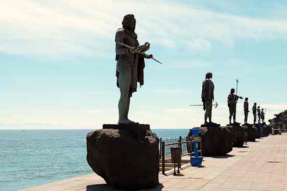 Bronsestatuer av mennesker med spyd står på rekke langs en kystpromenade. Figurene er plassert på steinsokler med hav og himmel som bakgrunn.