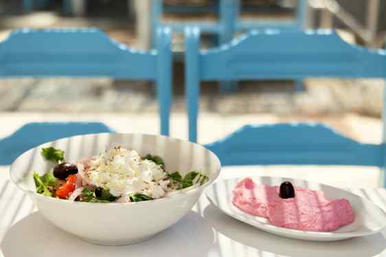 En salat med grønne blader og smuldret hvit ost serveres i en lys bolle. Ved siden av står en tallerken med rosa innhold, og blå stoler ses i bakgrunnen.