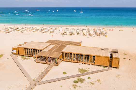 Flyfoto av sandstrand med solsenger, turkist hav og en moderne trebygning nær vannet