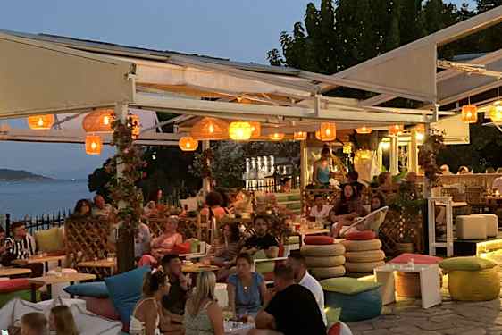 Livlig restaurant ved vannet med gjester, utendørs sitteplasser og hengende lamper under åpen pergola.