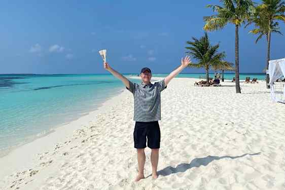 Glad Tomas på hvit sandstrand med turkisblått hav og palmer