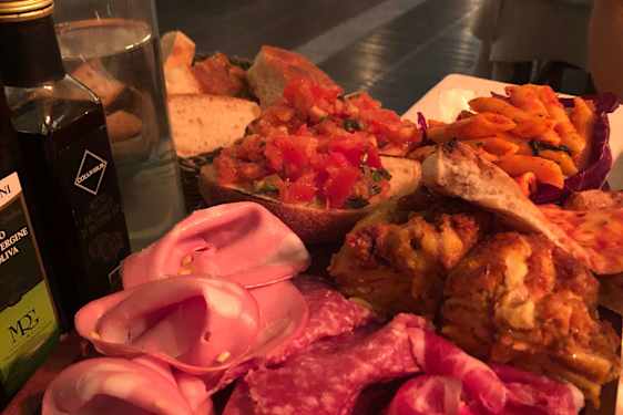 Antipastifat med spekemat, bruschetta, grillet kjøtt, pasta, brød og olivenolje servert på et restaurantbord.