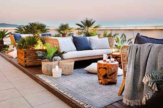 Et loungeområde på takterrassen er innredet med sofa, puter, tepper og grønne planter. Fra sitteplassen er det utsikt mot havet og den lyse horisonten.