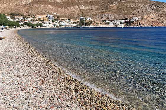 Klapperstensstrand med turkist hav, omgitt av fjell i horisonten og noen hus.