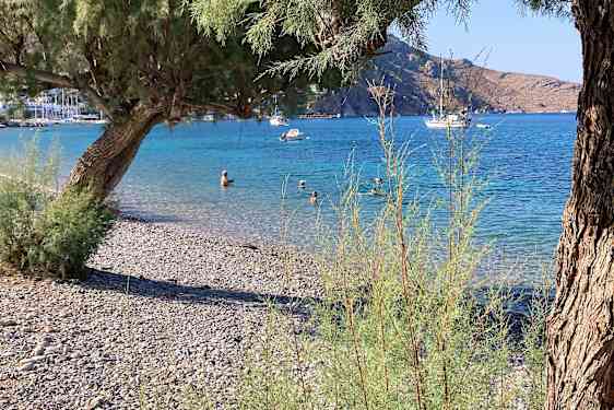 Klapperstensstrand med turkist hav, der noen bader og et par seilbåter ligger i vannet.