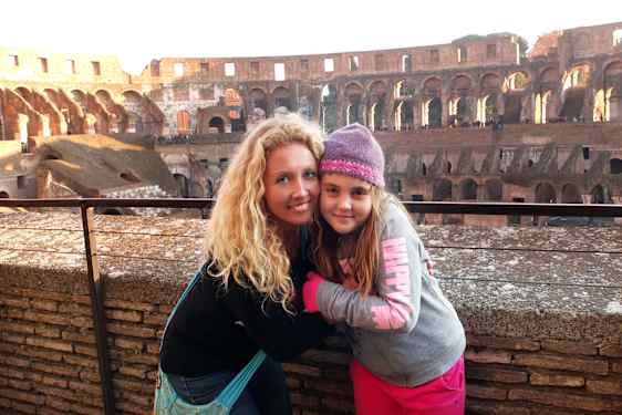 En kvinne med langt, lyst hår sitter tett sammen med en jente i grå genser og strikkelue. Colosseum fyller bakgrunnen.