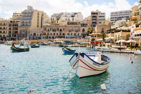 Bilde av båthavnen i St. Julian’s på Malta med fargerike fiskebåter, klart blått vann og historiske bygninger i bakgrunnen.