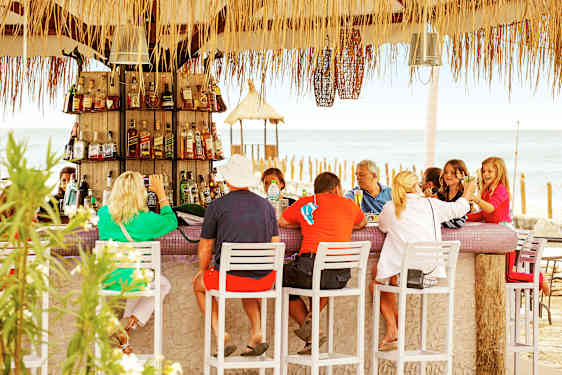 Flere voksne og barn sitter rundt en bar med hvite barkrakker. Flasker fyller hyllene bak disken, og havet er synlig i bakgrunnen.