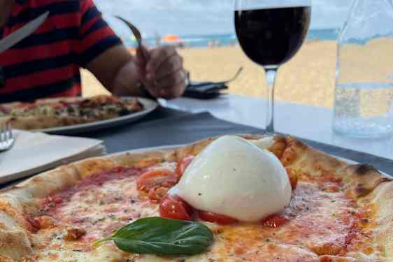 Nærbilde av en pizza med smeltet ost, røde tomater og frisk basilikum. En stor, rund kule hvit ost ligger på toppen, mens en person som spiser og en strand synes i bakgrunnen.