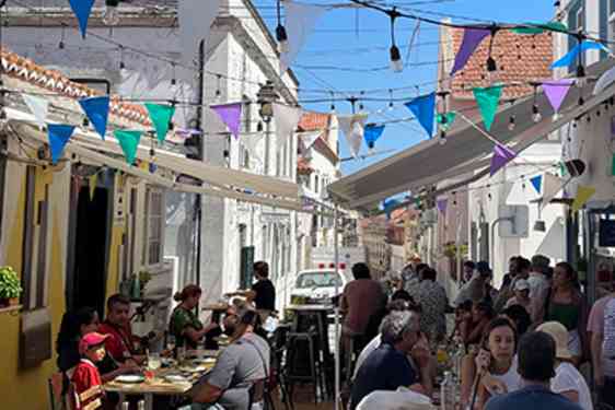 Fargerike vimpler henger over en livlig gate med restauranter og uteserveringer. Gjester sitter tett ved bordene mellom lyse bygninger under en skyfri himmel.