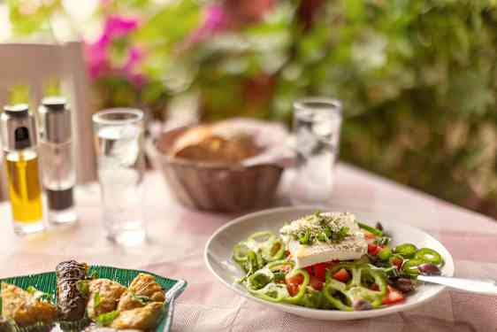 En tallerken med gresk salat står på et dekket bord med brød, glass og små flasker i bakgrunnen.