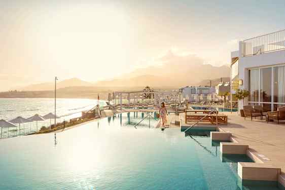 Hotell med stort basseng, havutsikt, hvite bygninger og fjell i bakgrunnen i varmt sollys.