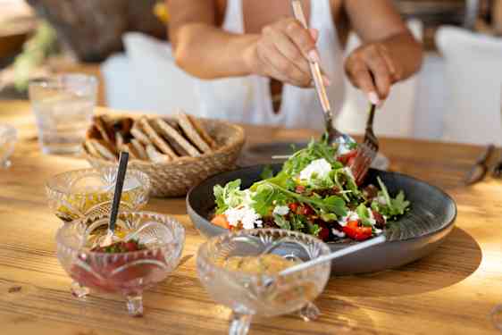 En stor skål med salat står på et trebord sammen med flere små skåler, en kurv med brød og et glass. En person forsyner seg av salaten med bestikk.