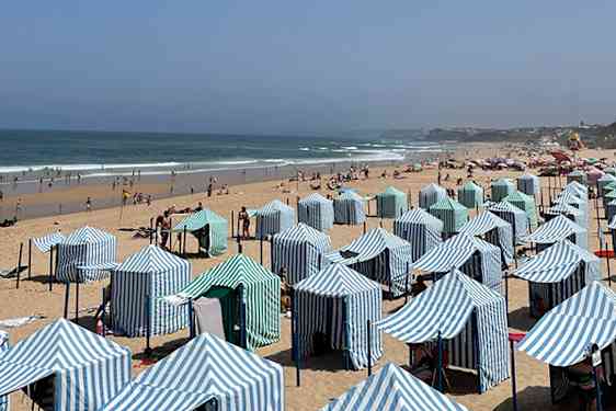 Rekker med blå- og hvitstripete strandtelt dekker den brede sandstranden. Badegjester nyter stranden ved det åpne havet under en klar, blå himmel.
