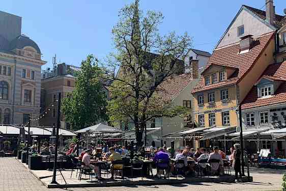 Uteserveringer med bord og parasoller fyller et åpent torg omgitt av bygninger. Trær og et grønt plenområde rammer inn plassen.