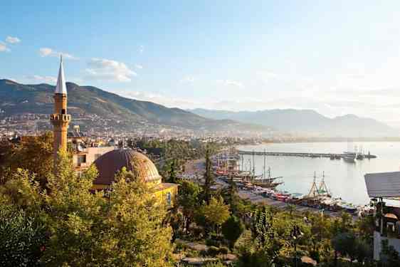 Et høyt, slankt tårn og en stor kuppel står mellom grønne trær. Nedenfor ligger en havn med mange båter, bebyggelse og fjell i bakgrunnen.