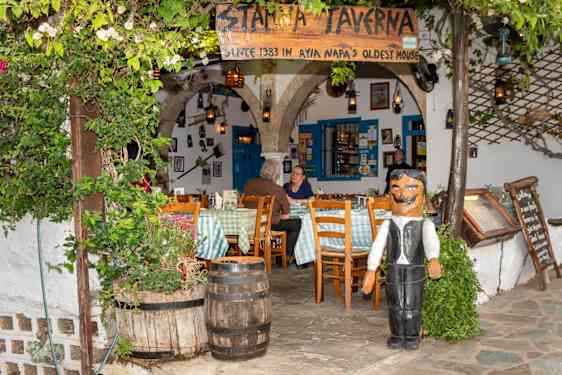 En restaurant med skiltet «Stamma Taverna» har hvite vegger, blå dører og buede åpninger. Bord, stoler, tønner, grønne planter og en trefigur av en servitør står utenfor.