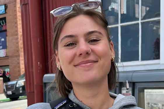 Portrett av en smilende person utendørs, med solbriller på hodet og en bygning i bakgrunnen.