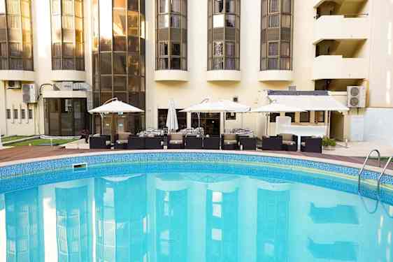 Det runde utendørsbassenget ligger foran hotellets terrasse med bord, stoler og parasoller. De lyse hotellbygningene speiler seg i det rolige vannet.