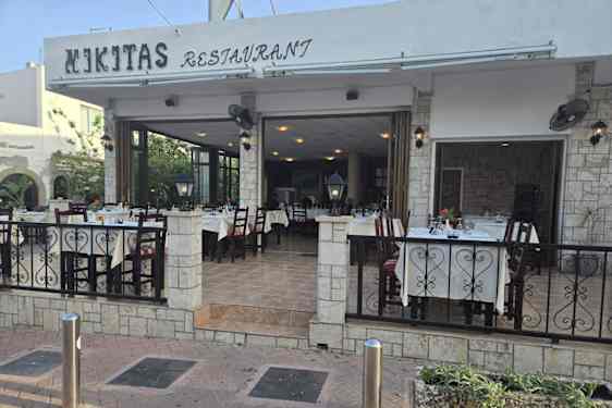 Bord og stoler står på en overbygd uteservering foran en åpen restaurant med navnet "Nikita restaurant" skreet på siden av taket. Området er avgrenset av lyse steinvegger og svart metallrekkverk.