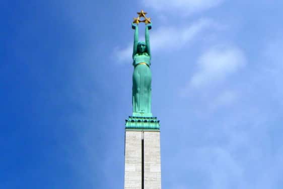 En slank søyle med lys stein bærer en stor grønn statue på toppen. Figuren løfter armene høyt over hodet og bærer tre stjerner, som troner mot den klare, blå himmelen.