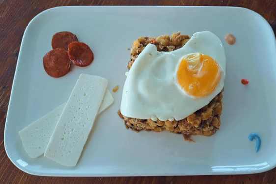En firkantet tallerken er dekket med en frokostvariant av cachupa, toppet med et speilegg og servert med pølsebiter og osteskiver.