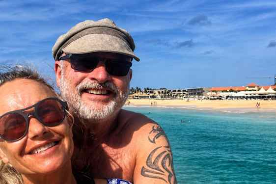 Et smilende par poserer foran klart, turkist vann og en lys sandstrand under blå himmel.